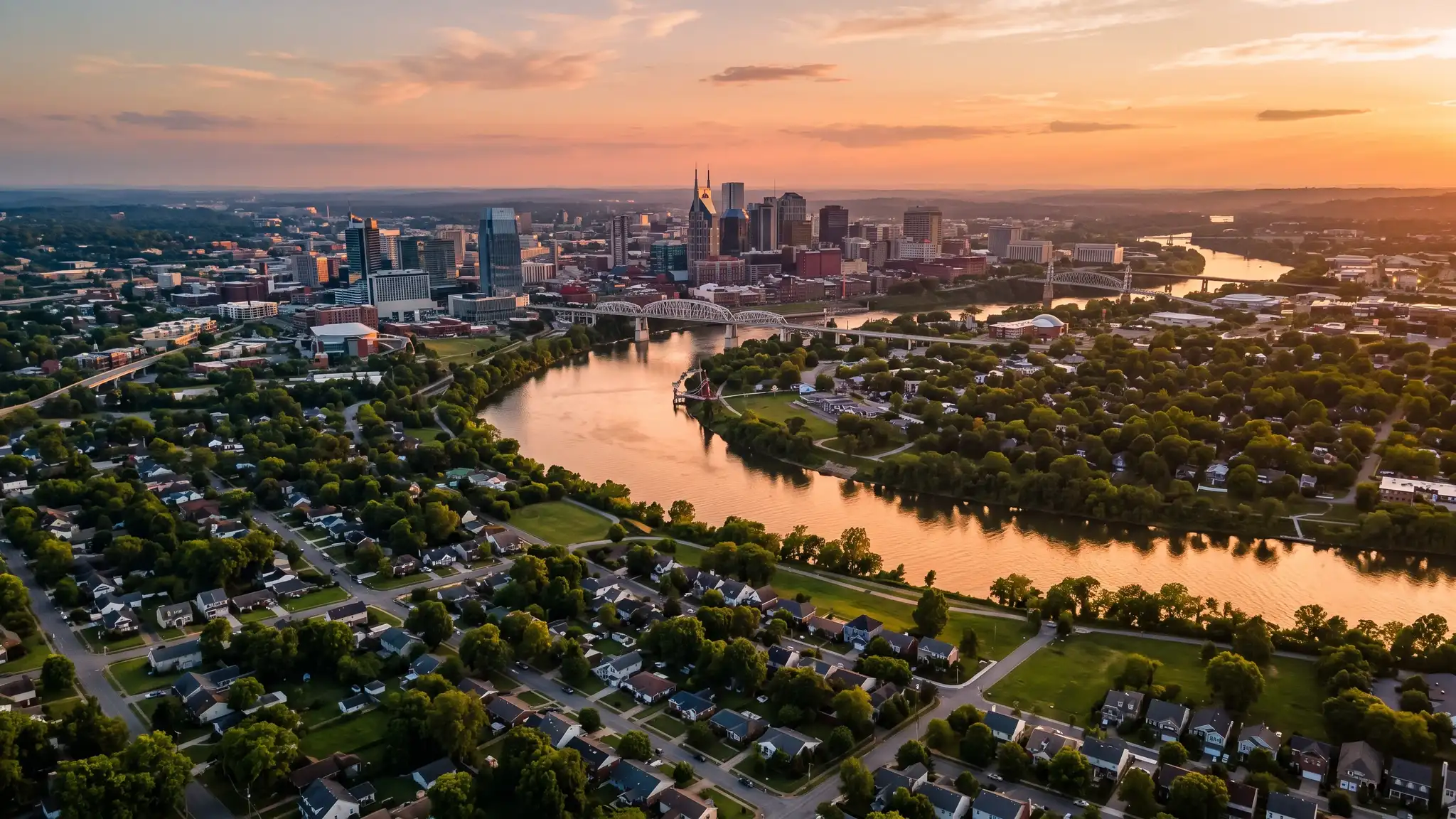 Aerial view of the Cumberland River winding past the Nashville skyline and residential neighborhoods at sunset with golden light on the water